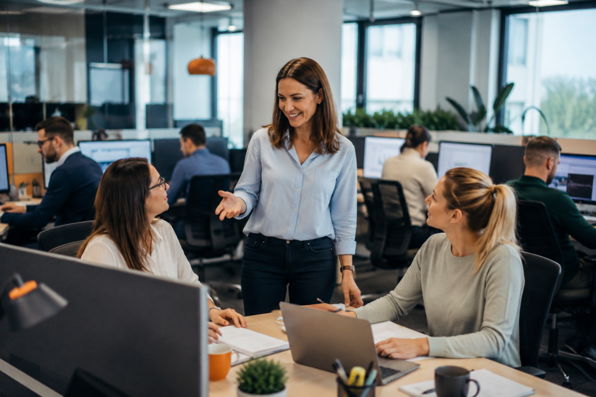 Three colleagues collaborating in a busy modern open-plan office while other team members work in the background.