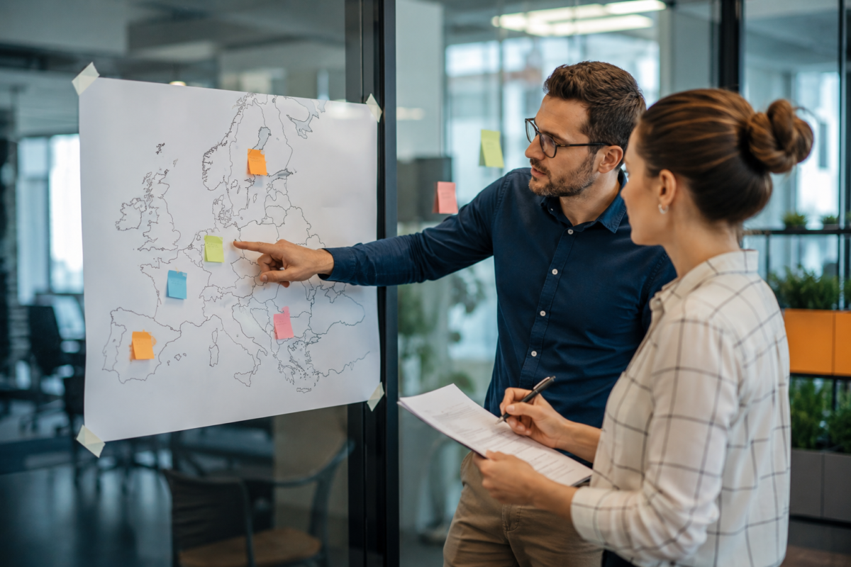 Two colleagues reviewing a map of Europe with sticky notes while cross-checking a printed document in a modern office.